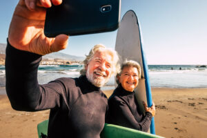 älterer Herr macht ein Selfie von sich und einer älteren Dame, die beide mit Surfbrettern am Strand vor dem Meer stehen
