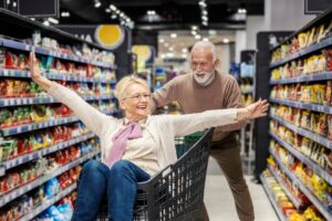 älterer Herr schiebt ältere Frau im Einkaufswagen durch Supermarkt-Gänge, beide sehen glücklich aus.