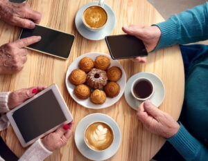 Draufsicht auf einen Tisch an dem ältere Hände Smartgeräte halten und auf dem Tassen mit Kaffee und Tee und ein Teller italienischer Kuchen stehen<br />